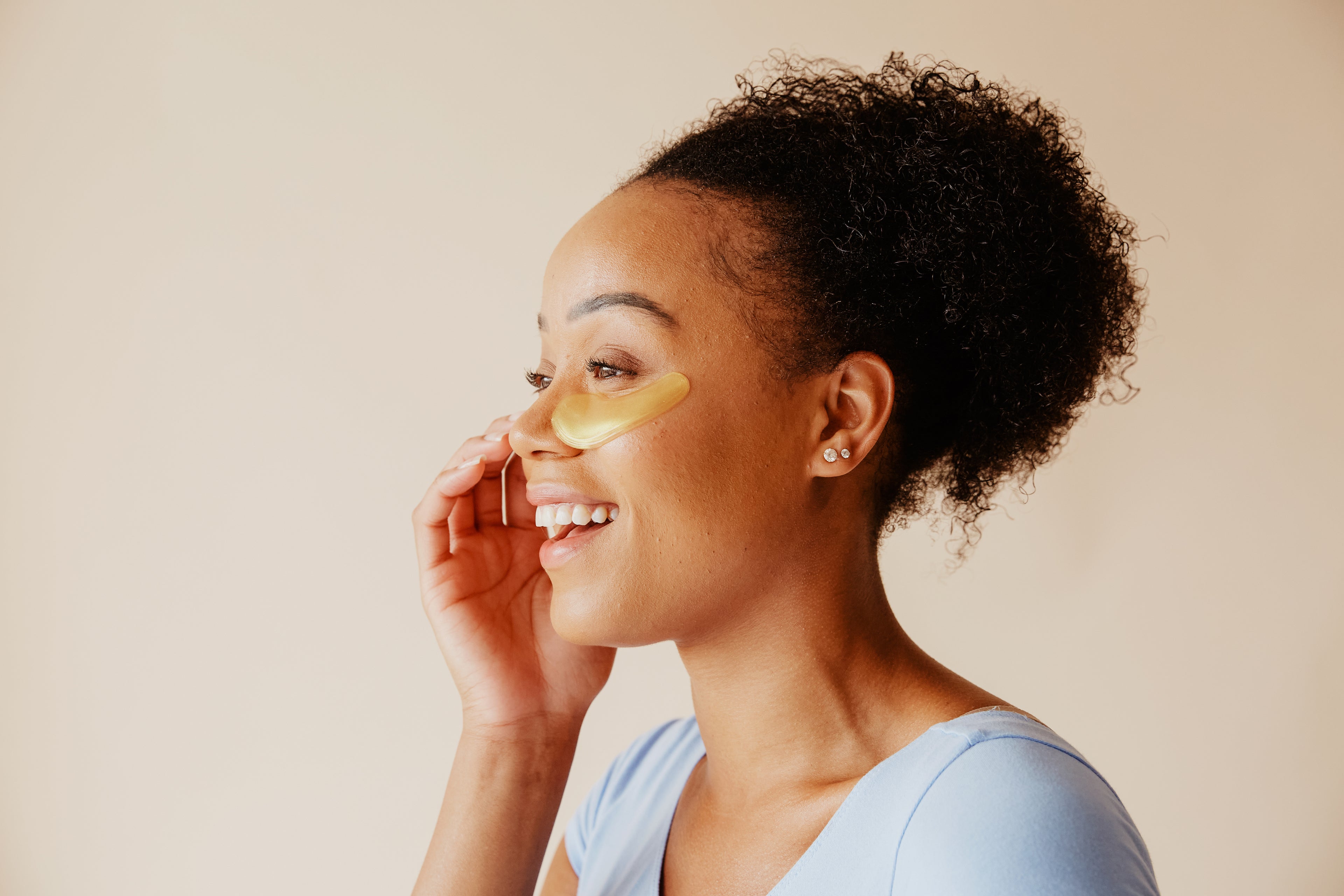 Smiling woman applying under-eye patches for skincare on a light background.